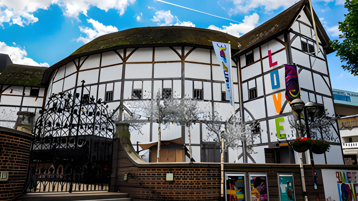 Shakespeare's Globe Theatre in London, England. It is a building with windows, a metal fence, a signboard, a street pole, a group of trees, some plants in pots, a banner with some text on it, some poles, a wire, and a cloudy sky.
