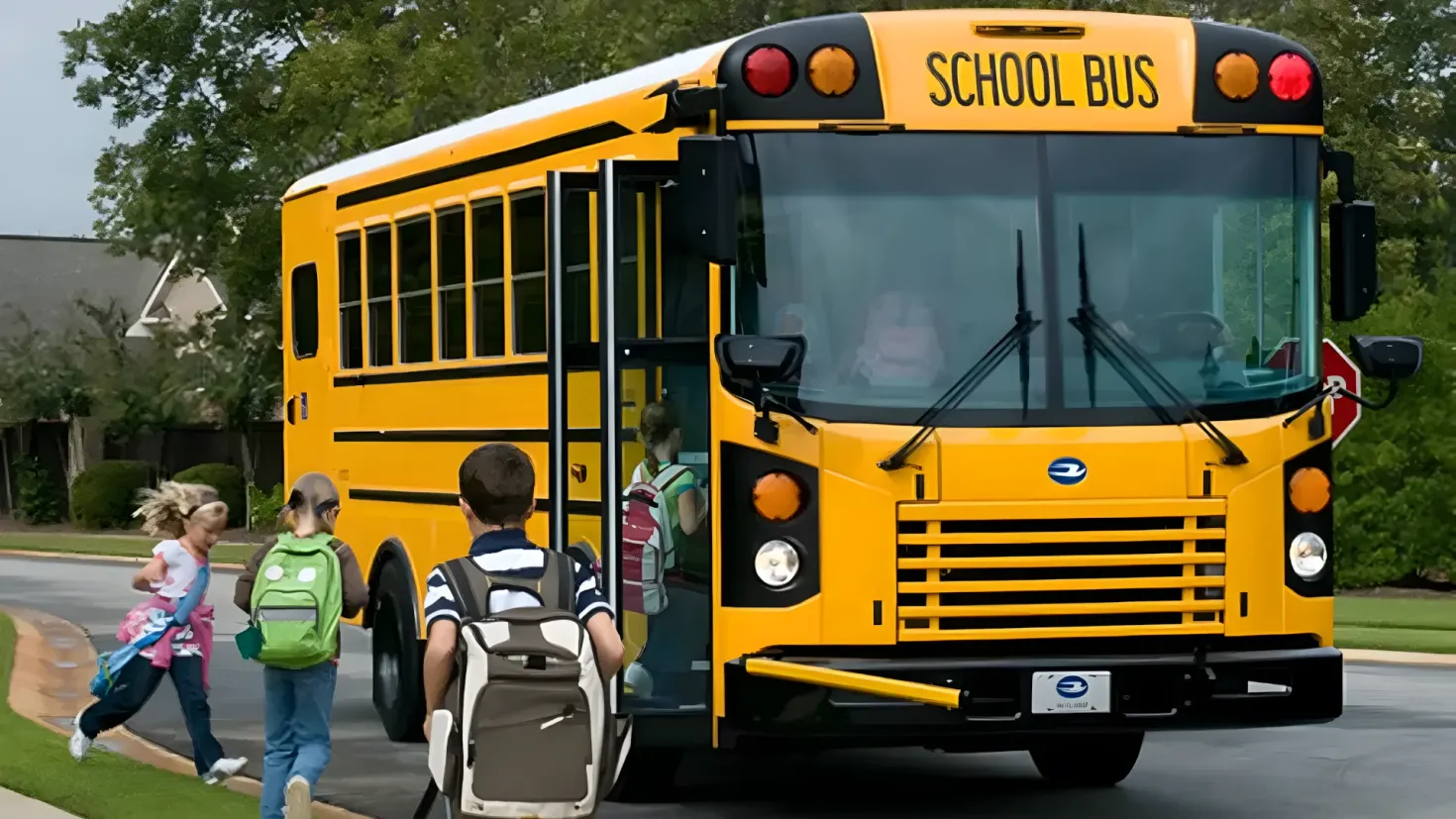A yellow school bus driving down a street with a group of children walking towards it. The children are wearing backpacks and one of them is holding a stroller. The street is lined with grass, plants, trees, and houses, and the sky is visible in the background.