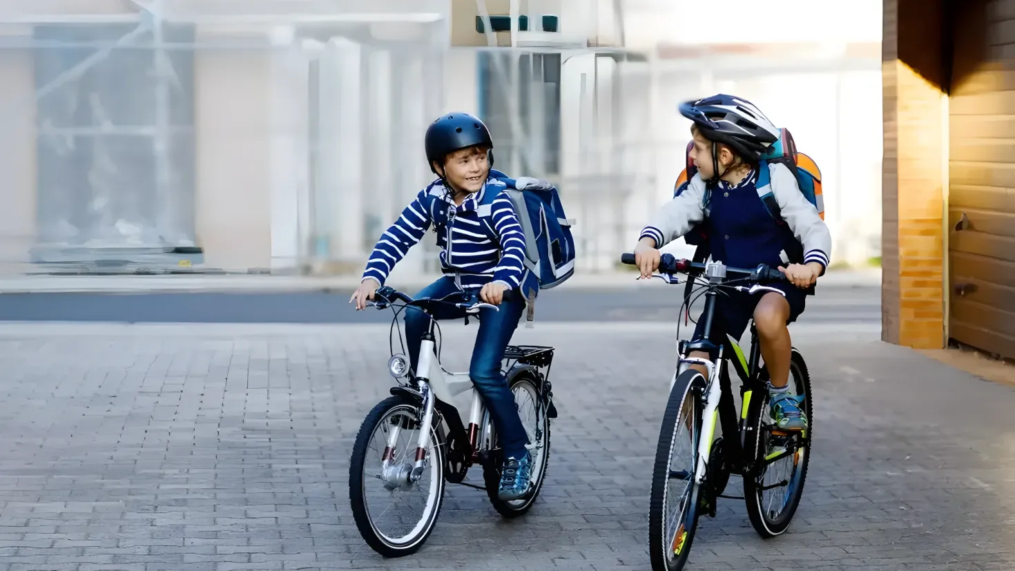 Two young boys riding bicycles down a street, both wearing helmets and carrying backpacks. The background is slightly blurred, giving the image a sense of motion.