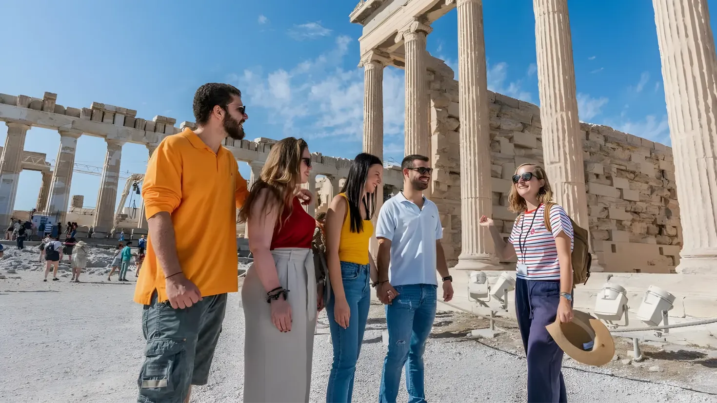 A group of people standing in front of the Parthenon temple in Athens, Greece. Some of the people are wearing goggles and one person is wearing a hat. The sky is filled with clouds and the temple is surrounded by pillars.