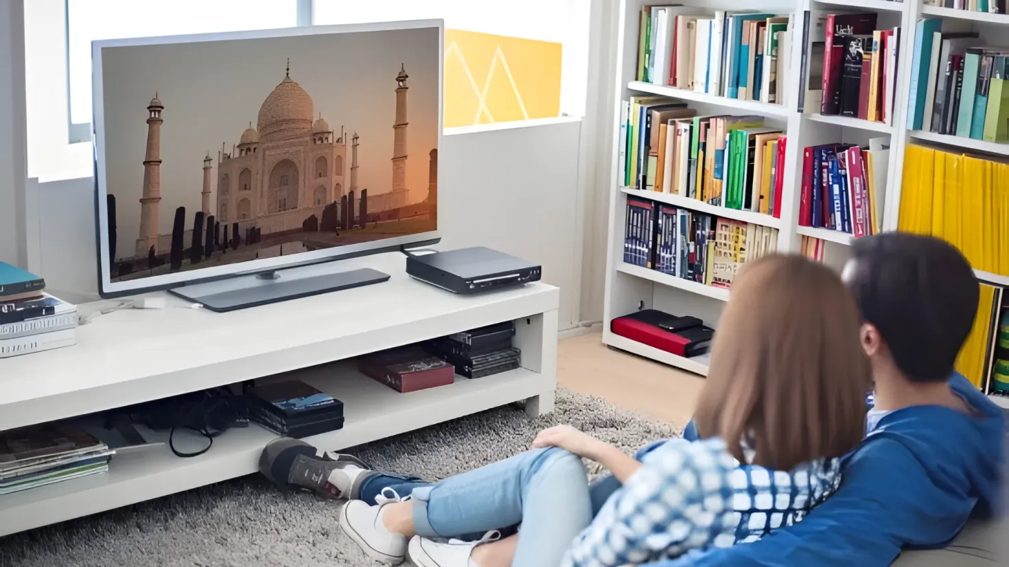 A man and woman sitting on a couch in front of a television, surrounded by bookshelves filled with books. On the television screen, there is an image of the Taj Mahal, and on the table beside it, there are books, a device, and other objects. The floor is covered with a carpet, and there is a window in the background.