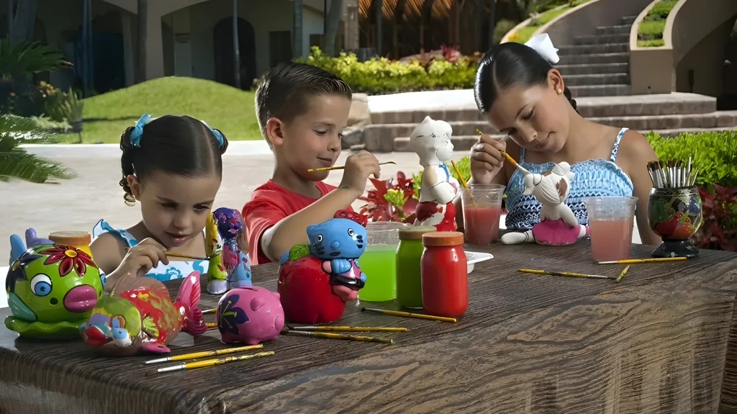 Three children sitting at a table with a variety of toys, brushes, glasses, and other objects. In the background, there are plants, trees, steps, and a building.