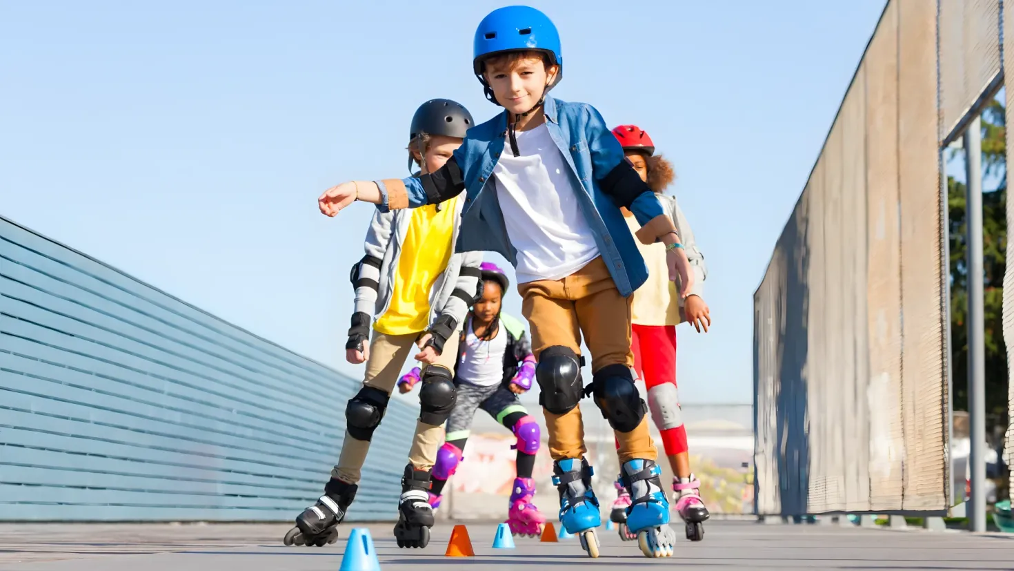 A group of children rollerblading down a sidewalk, wearing helmets, knee pads, and skating shoes. The sidewalk is lined with traffic cones and there is a wall on the right side of the image. In the background, there are trees and a clear blue sky.