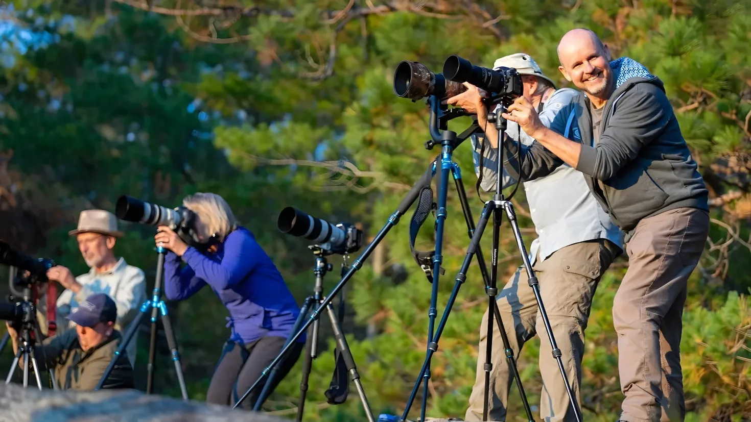 A group of people standing next to each other with cameras on stands, surrounded by trees and a clear blue sky. Some of the people are wearing hats, suggesting they are taking pictures.