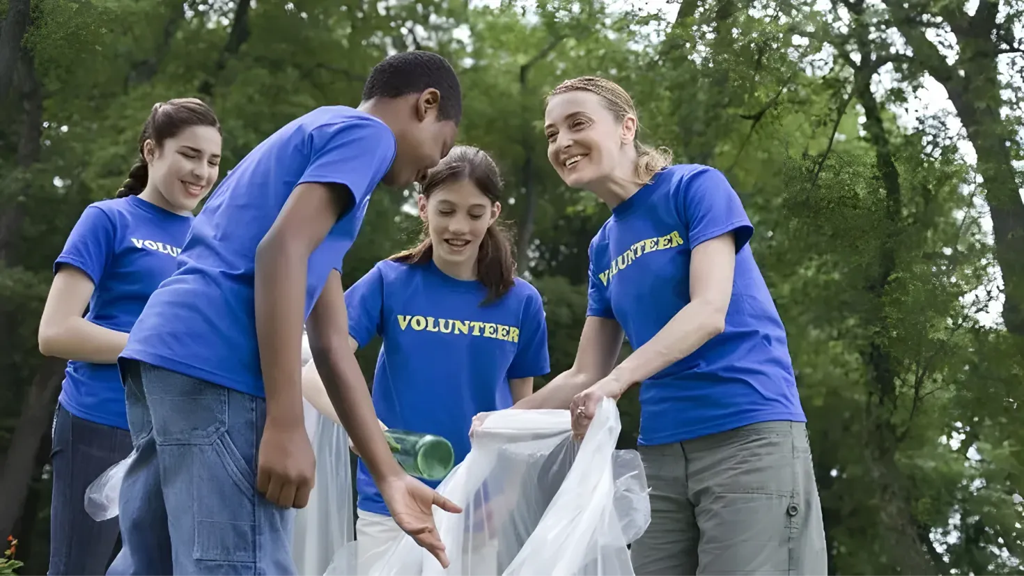 A group of four people in blue t-shirts standing next to each other, each holding a plastic cover and a bottle. In the background, there are trees and a clear blue sky.