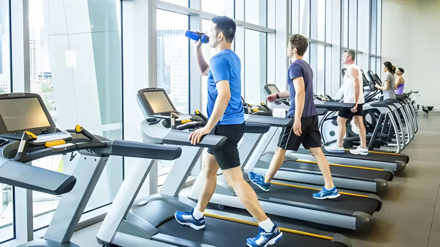 A group of people running on treadmills in a gym, with a wall on the right side and glass windows on the left side, through which buildings and the sky can be seen.