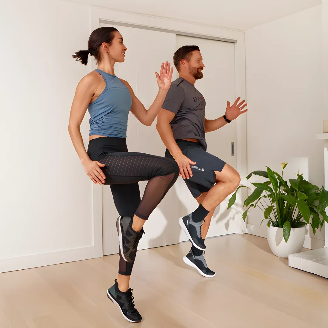 A man and woman doing squats in a living room. The room is decorated with a plant in a pot, some objects placed on the shelves, a door, and a roof with ceiling lights.