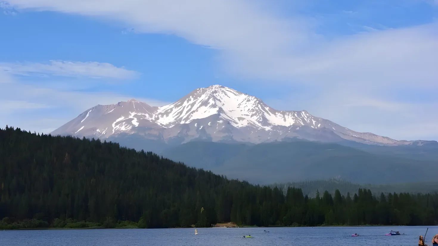 A group of people in kayaks paddling on a lake with a majestic mountain in the background. The lake is surrounded by lush green trees and the sky is filled with white, fluffy clouds.
