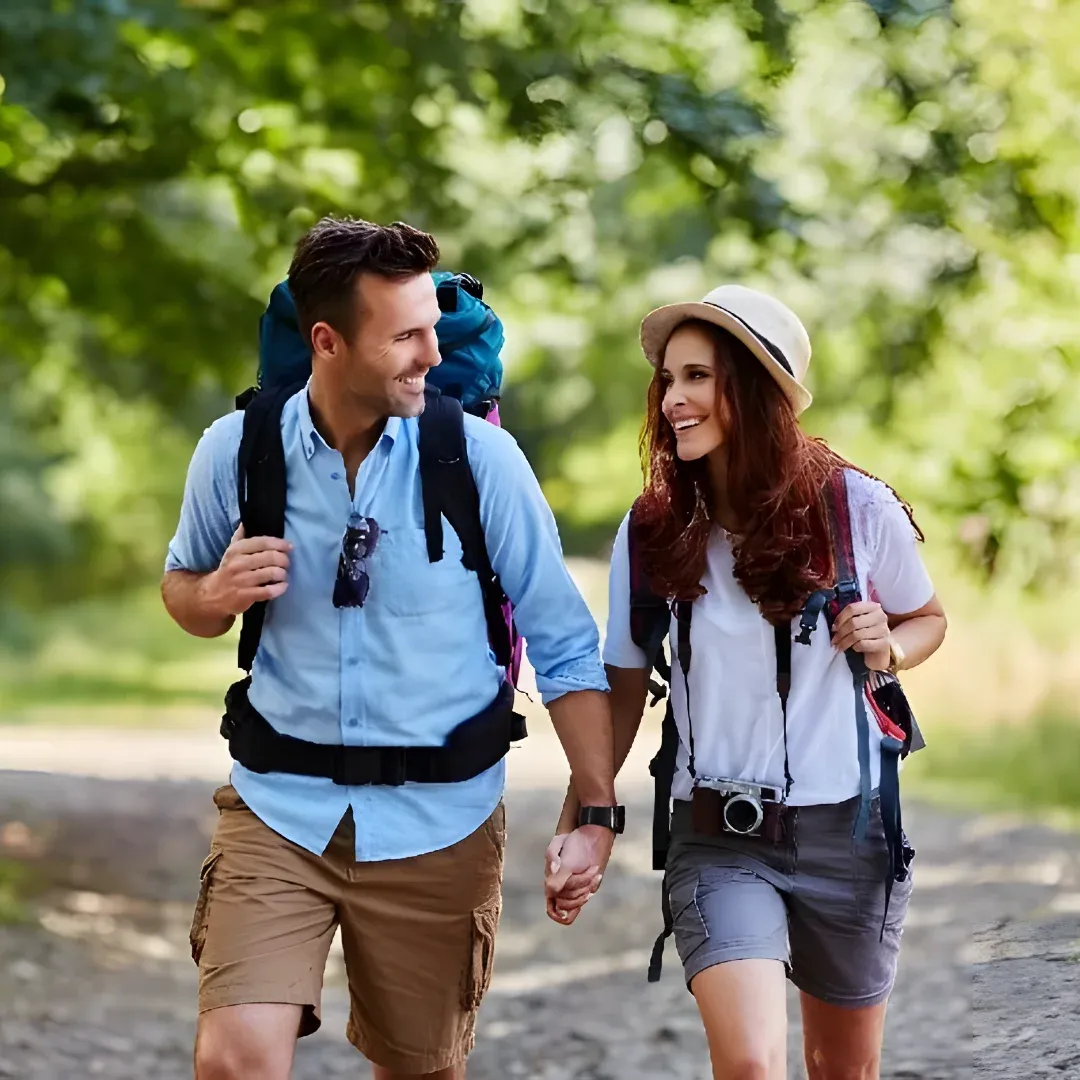 A man and woman walking down a sidewalk with backpacks on their backs. The man is wearing a blue and white shirt and cream pants, while the woman is carrying a blue, red, and black backpack with a solar panel attached to it. In the background, there are a few trees in green and a white building.