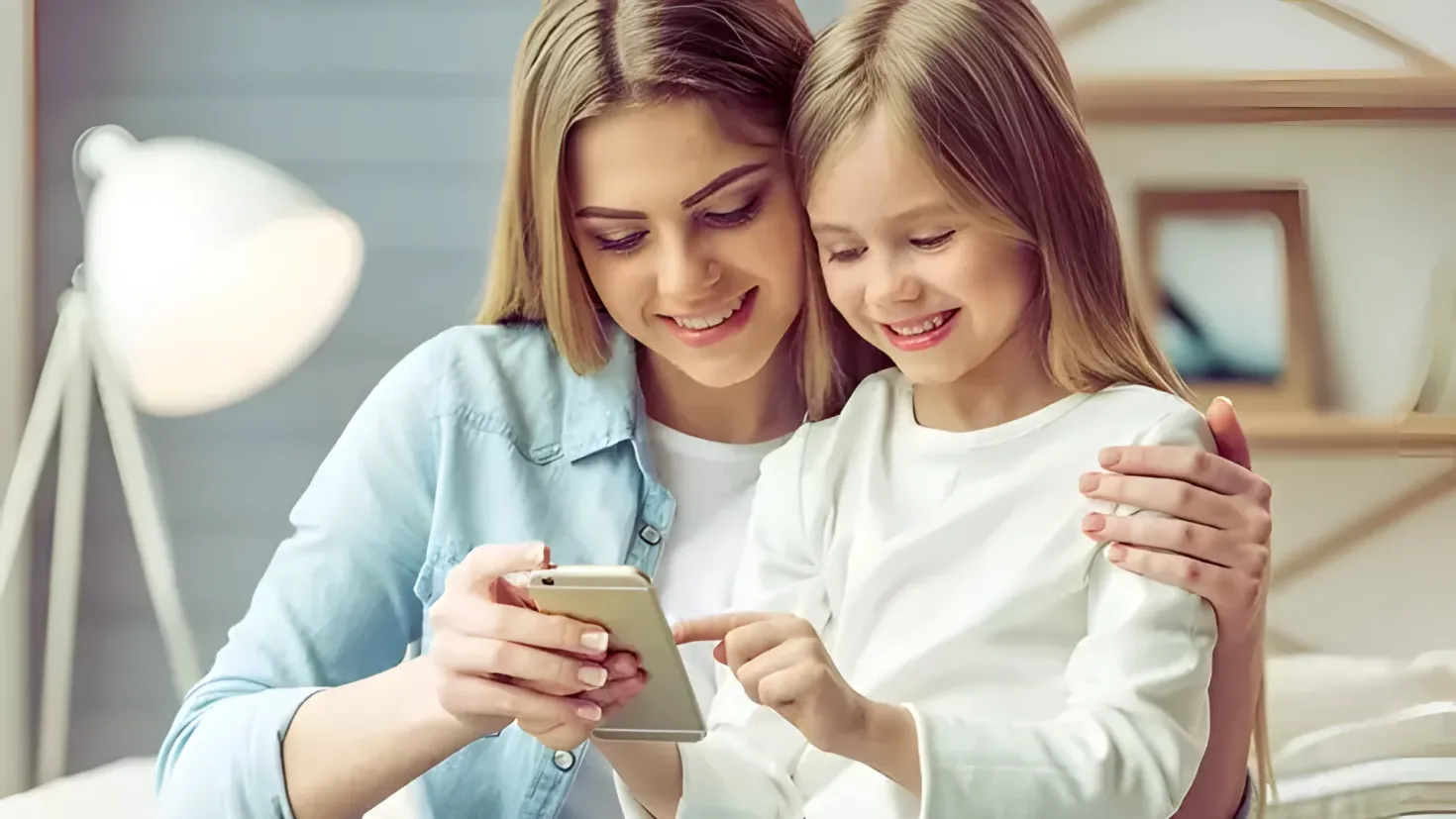 A mother and daughter sitting on a bed, both of them looking at a cell phone. The mother is holding the phone in her hands, while the daughter is looking at it intently. In the background, there is a lamp, a frame on the wall, and a hanger.