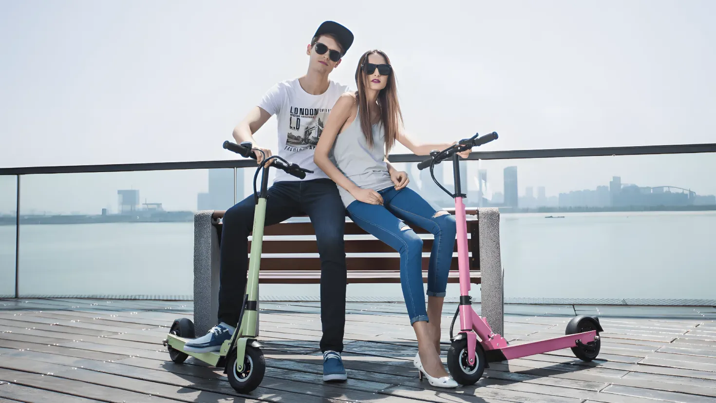 A man and woman sitting on a bench next to each other, both wearing white t-shirts, blue jeans, and black shoes. Behind them is a railing, and in the background there is a body of water, buildings, and a clear blue sky.