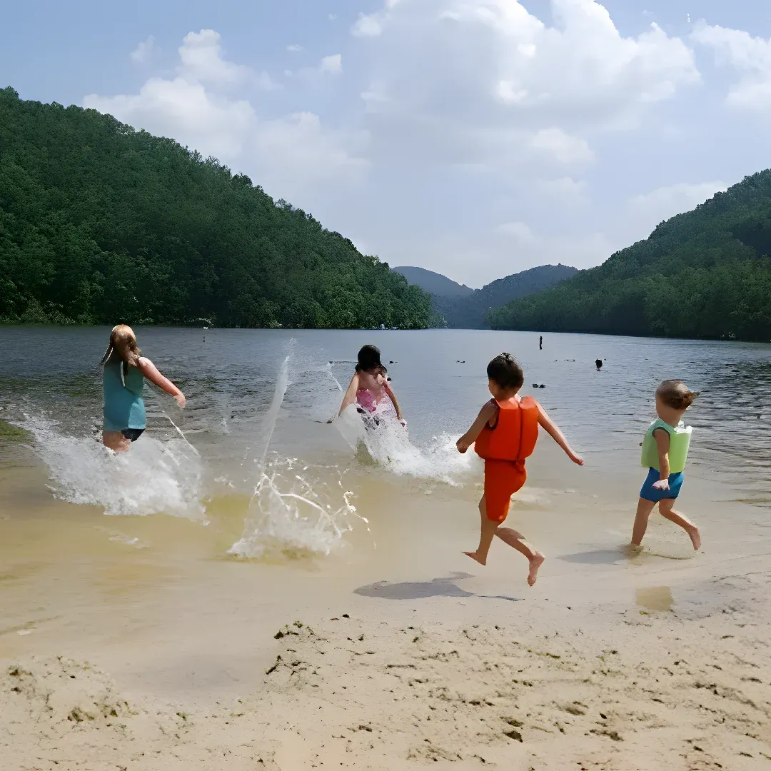 A group of children playing in the water at the shore of a lake, with sand beneath their feet and trees and hills in the background. The sky is filled with clouds, creating a peaceful atmosphere.