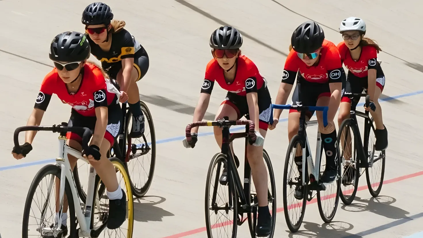 A group of people wearing helmets, goggles, gloves, and shoes riding bicycles down a street.