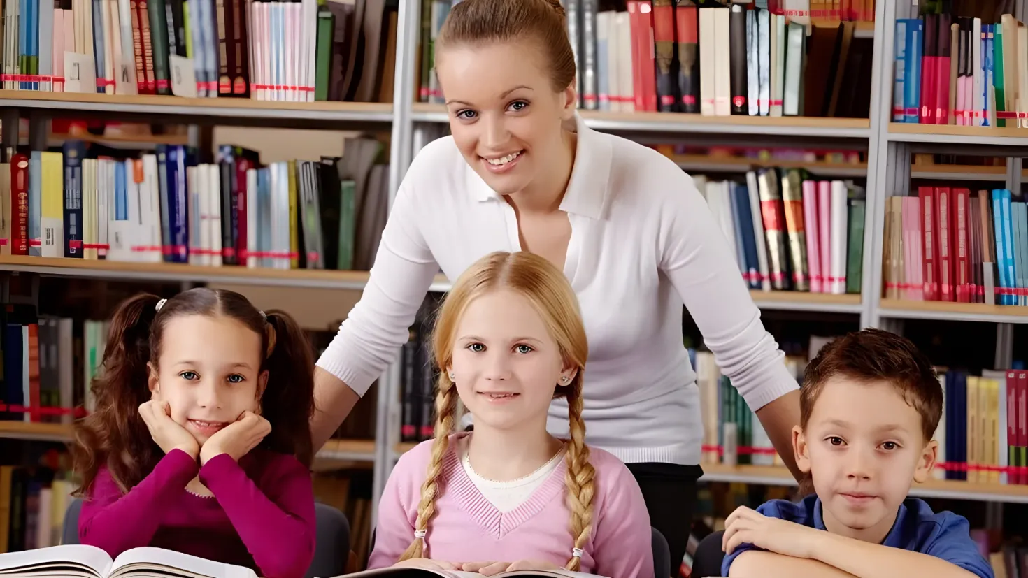 A teacher standing in front of a group of children in a library. The children are sitting on chairs and reading books, while the teacher is standing behind them. In the background, there are bookshelves filled with books.
