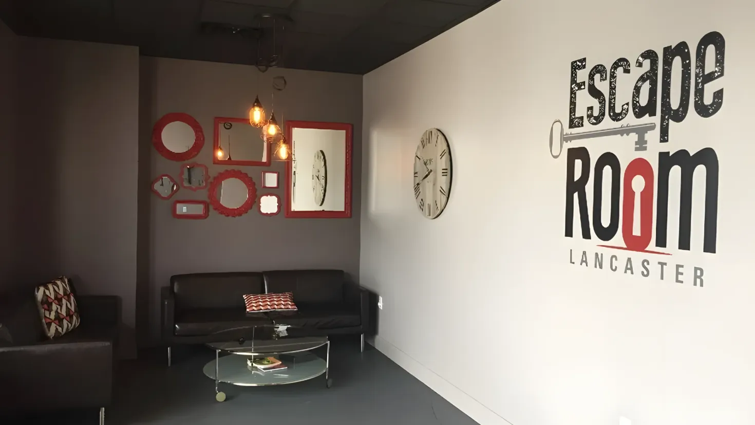 An escape room in Lancaster, with a studio couch, a coffee table, pillows, frames, a clock, and lights hanging from the ceiling. On the right side of the image, there is some text.