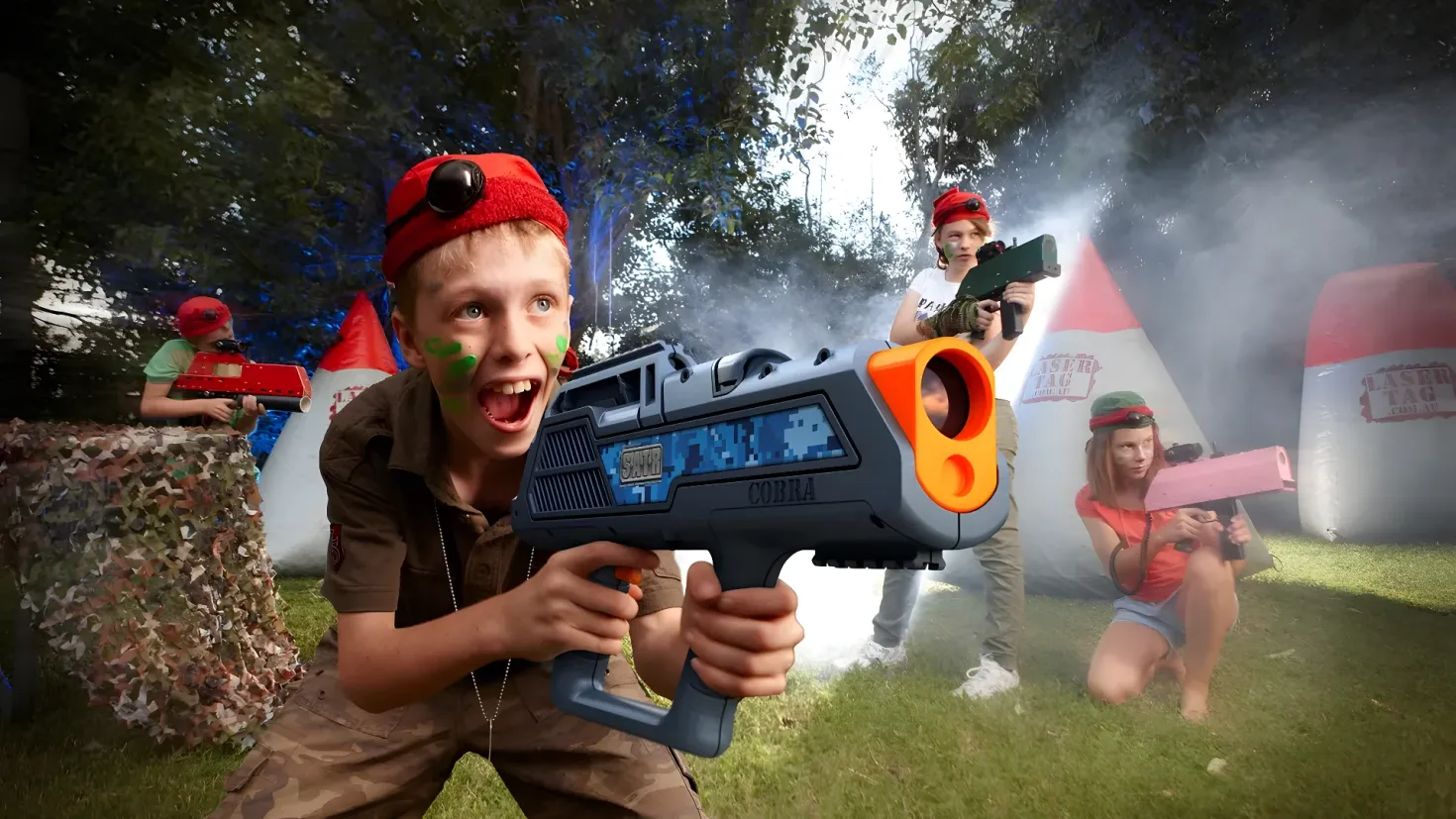 A group of children playing with Nerf guns in a grassy area surrounded by trees and a clear blue sky. One of the children is holding a toy gun in their hands.