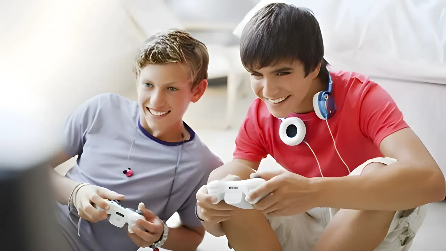 Two young boys sitting on the floor, each holding a video game controller in their hands and wearing headphones. The background is slightly blurred, suggesting they are focused on the game they are playing.