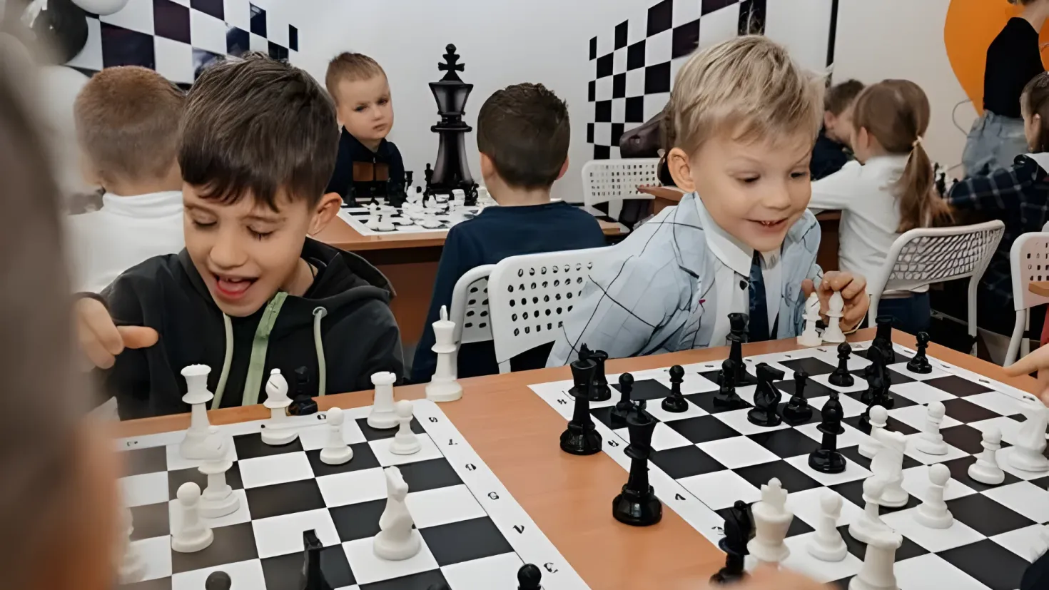 A group of children playing chess in a room. There are chess boards and pieces on the table, and a person standing in the background. The room is decorated with balloons and a wall.