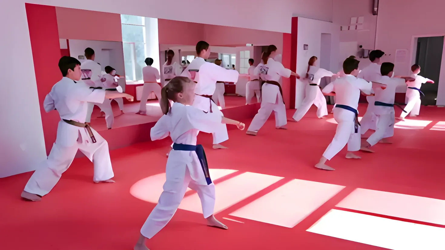 A group of people in karate gear practicing karate in a room with a red floor, walls, windows, doors, and other objects in the background. There is also a person wearing a belt in the center of the group.