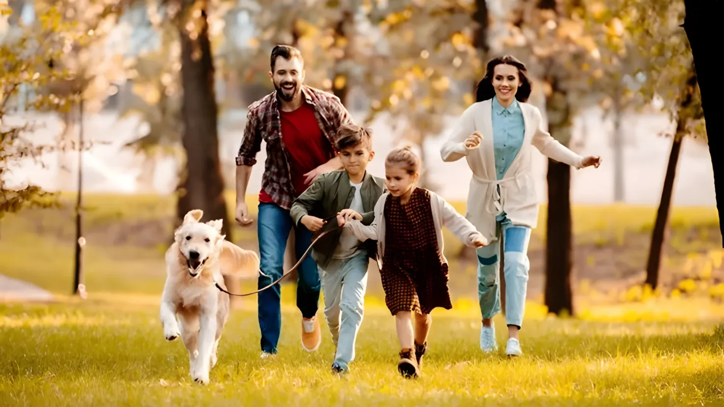 A happy family of four walking their dog in the park. The man and woman are holding the dog's leash, while the two children are running alongside them. The background of the image is filled with lush green grass, tall trees, and a bright blue sky.