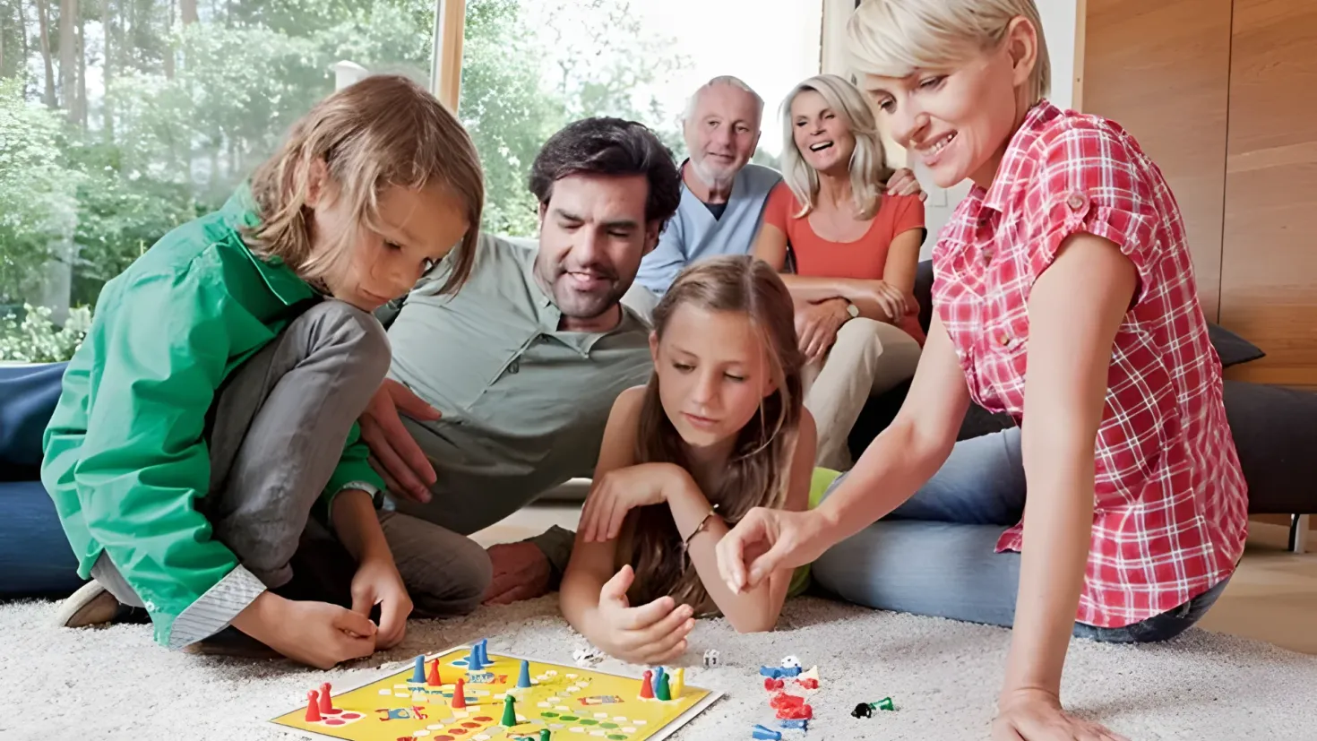 A family of four sitting on the floor playing a board game. The game board is filled with colorful pieces and the family is gathered around it. In the background, there is a sofa with pillows, a wooden cupboard, and a glass window with trees visible outside.