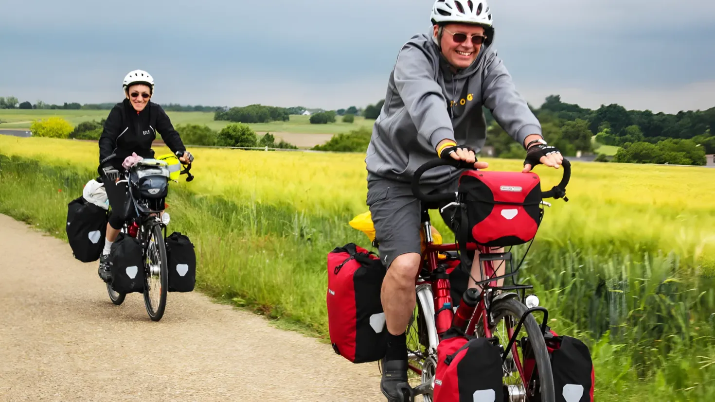 Two people riding bicycles down a dirt road surrounded by lush green grass and trees, with a bright blue sky in the background. They are both wearing helmets and carrying bags, suggesting they are on a bike tour.