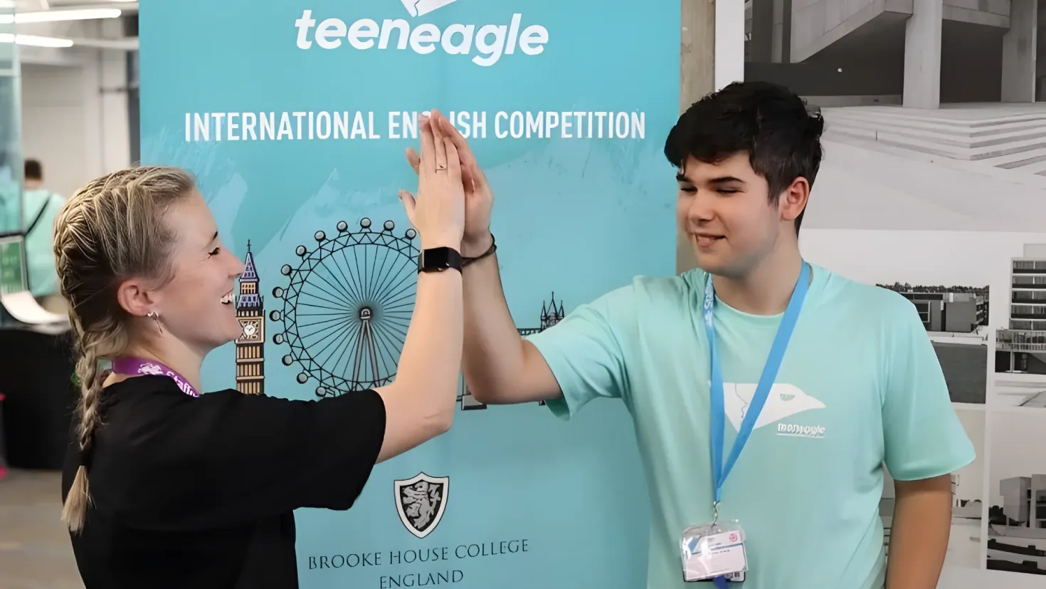 A man and woman standing next to each other in front of a sign that reads "Teenage International English Competition". The woman is wearing a black dress and the man is wearing an ID card. In the background, there are a few people, lights, and other objects.
