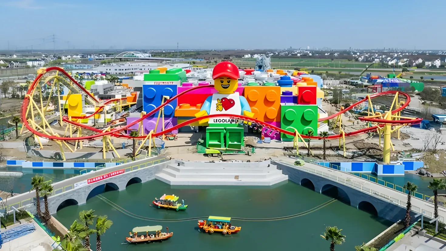 An aerial view of a Lego roller coaster in the middle of a body of water, surrounded by trees, buildings, and a bridge. There are also a few boats on the water, and the sky is visible at the top of the image.