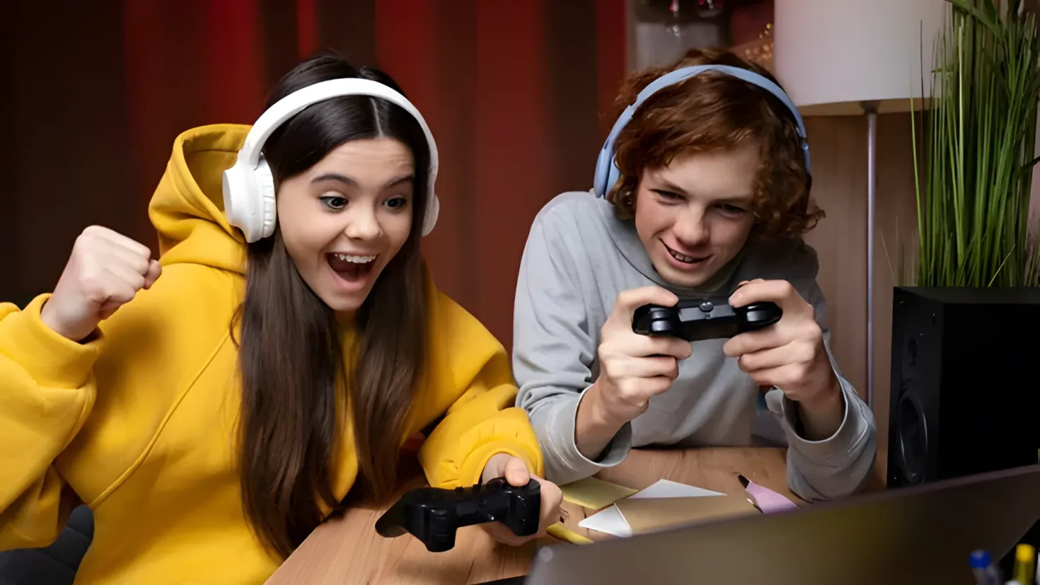 Two people sitting at a table, both wearing headphones and holding joysticks in their hands. On the table in front of them is a laptop, papers, pens, and other objects. To the right of the table is a plant and a lamp, and in the background is a red curtain.
