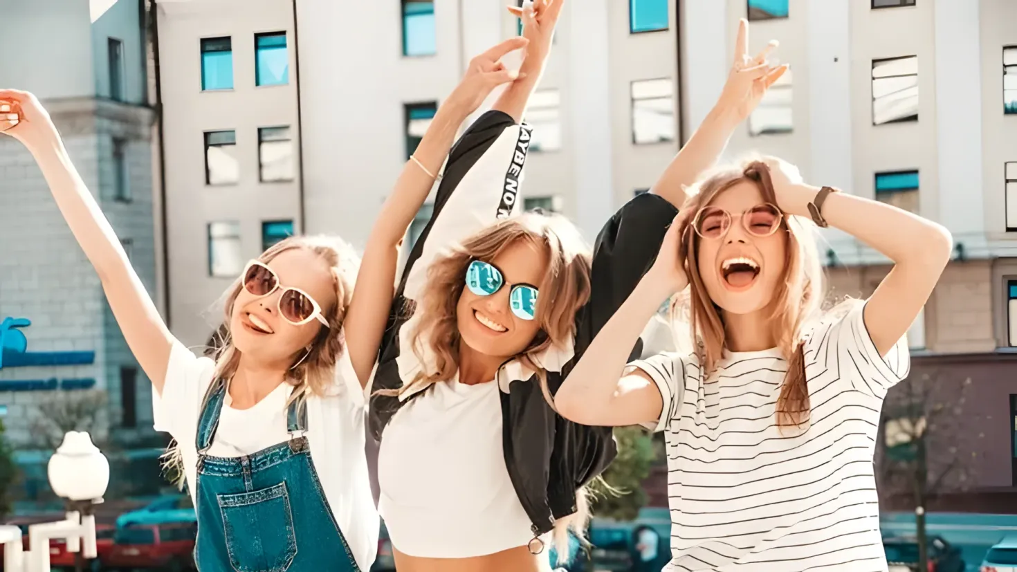 Three young women wearing sunglasses and posing for a photo in front of a building. In the background, there are vehicles on the road, light poles, trees, and buildings with windows.