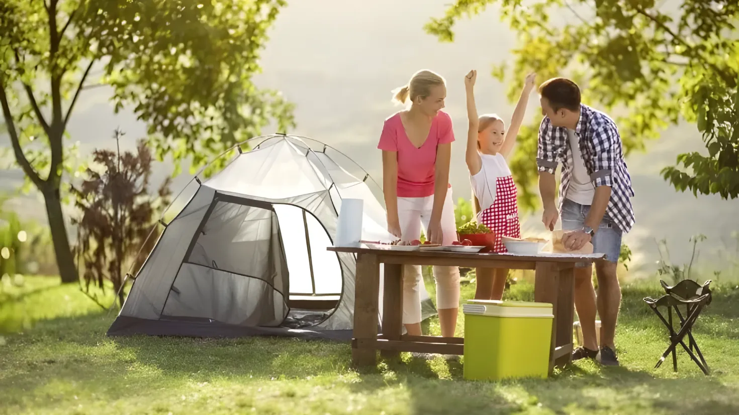 A family of four - a man, a woman and two children - standing around a picnic table in front of a tent. On the table there are plates, bowls, and other objects, and there is a box and a chair nearby. The ground is covered in grass and there are trees in the background, with a clear blue sky above.