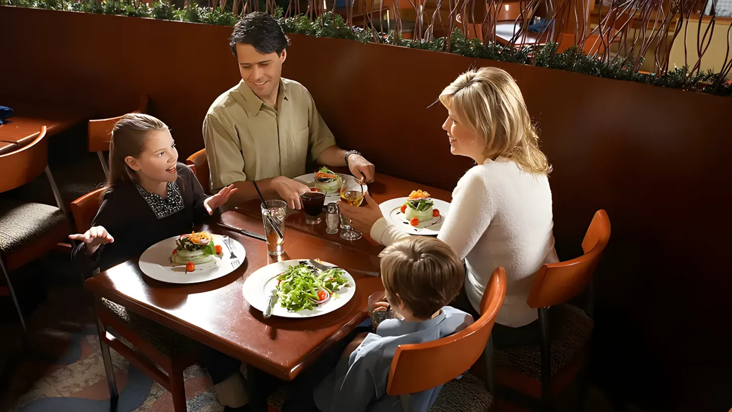 A family of four - a man, woman and two children - sitting around a table in a restaurant. On the table are plates of food, glasses, spoons, forks, knives, and other objects. In the background, there are plants, chairs, and a wall.