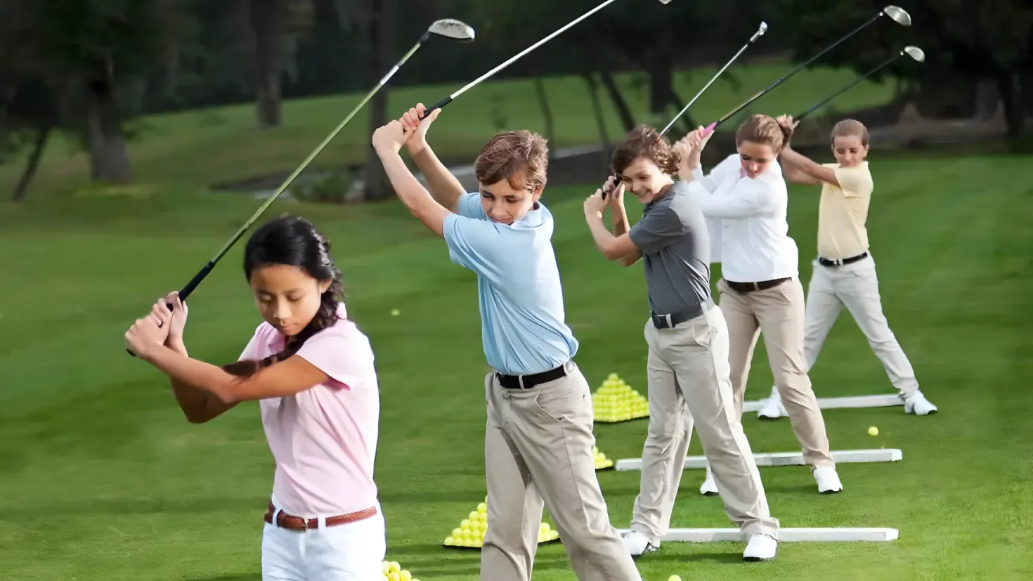 A group of people playing golf on a lush green field, with trees in the background. They are all holding golf clubs and there are golf balls scattered around them.