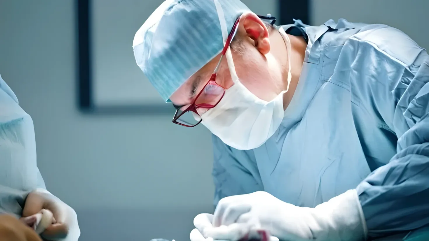 Two surgeons in scrubs and masks performing surgery on a patient in an operating room. The surgeon on the left is wearing a cap, glasses, and gloves, and is holding an object in his hand. The background is blurred, suggesting the focus is on the surgeons and the patient.