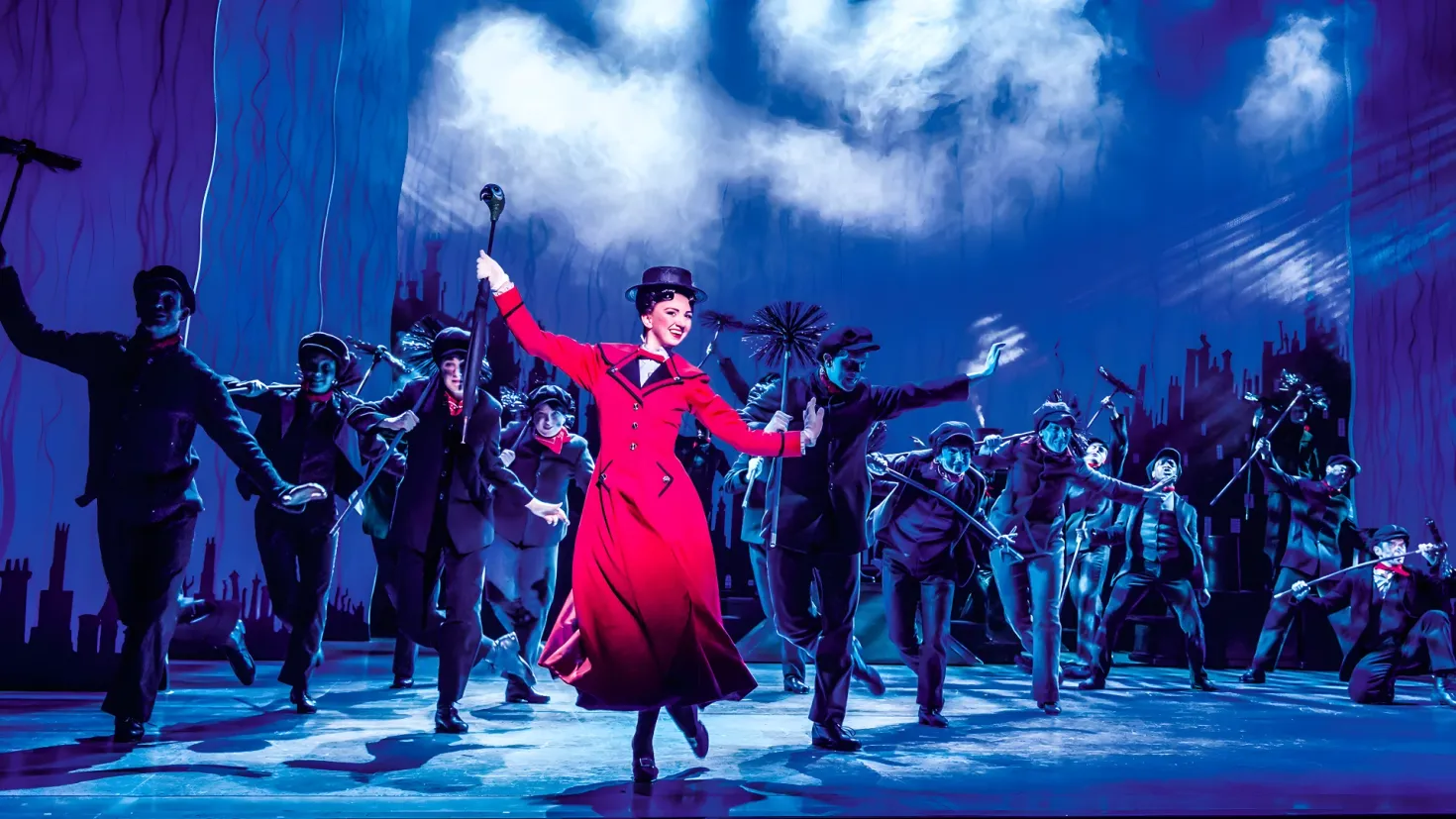 A woman in a red dress and hat dancing on a stage, surrounded by a group of people wearing costumes and holding various objects. In the background, there is a screen with buildings and a sky with clouds, creating a magical atmosphere reminiscent of the musical 'The Phantom of the Opera'.