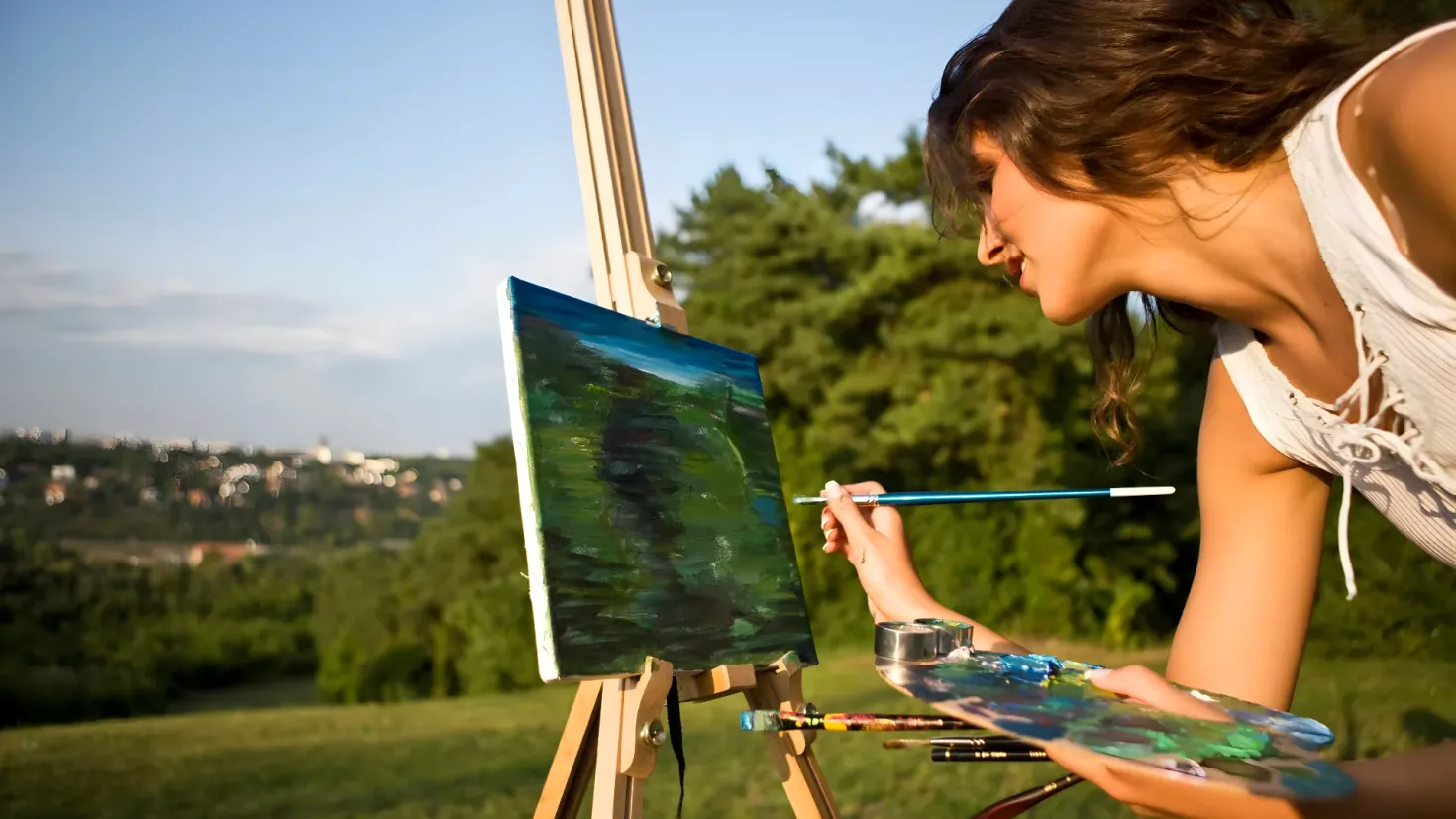 A woman painting on an easel in a park, surrounded by trees, buildings, and a clear blue sky. She is holding a paintbrush and a palette in her hands, and there are several other paintbrushes scattered around her.