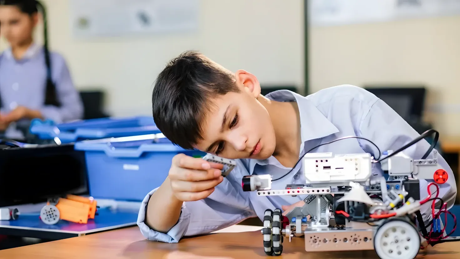 A young boy sitting at a table with a robot in front of him. On the table there are various objects, and in the background there is another person sitting on a chair. The background is slightly blurred, suggesting that the focus of the image is on the boy and the robot.