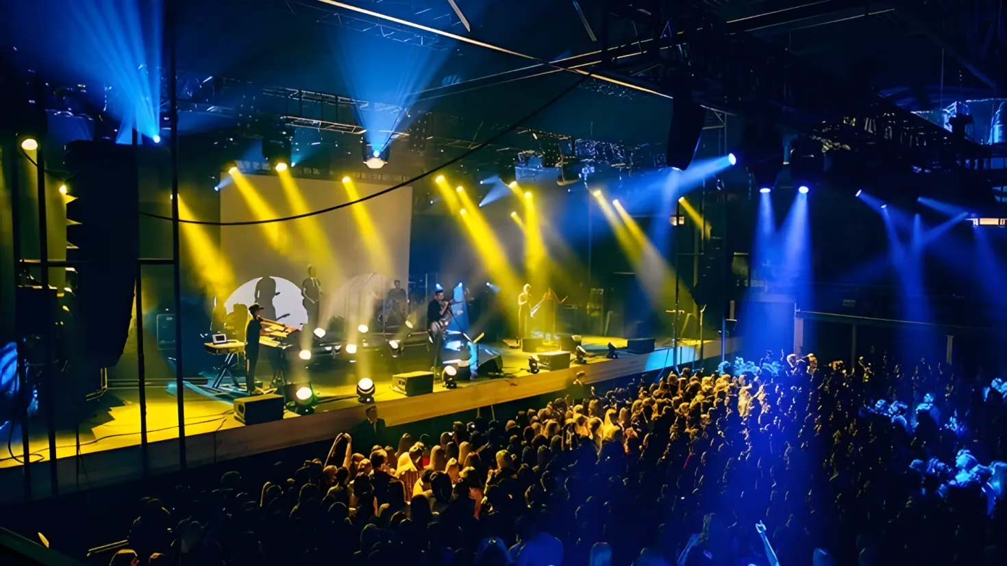 A large crowd of people standing in front of a stage, illuminated by bright lights. On the stage, a few people are playing musical instruments, and there is a loudspeaker in the foreground. The background is dark, creating a dramatic atmosphere.