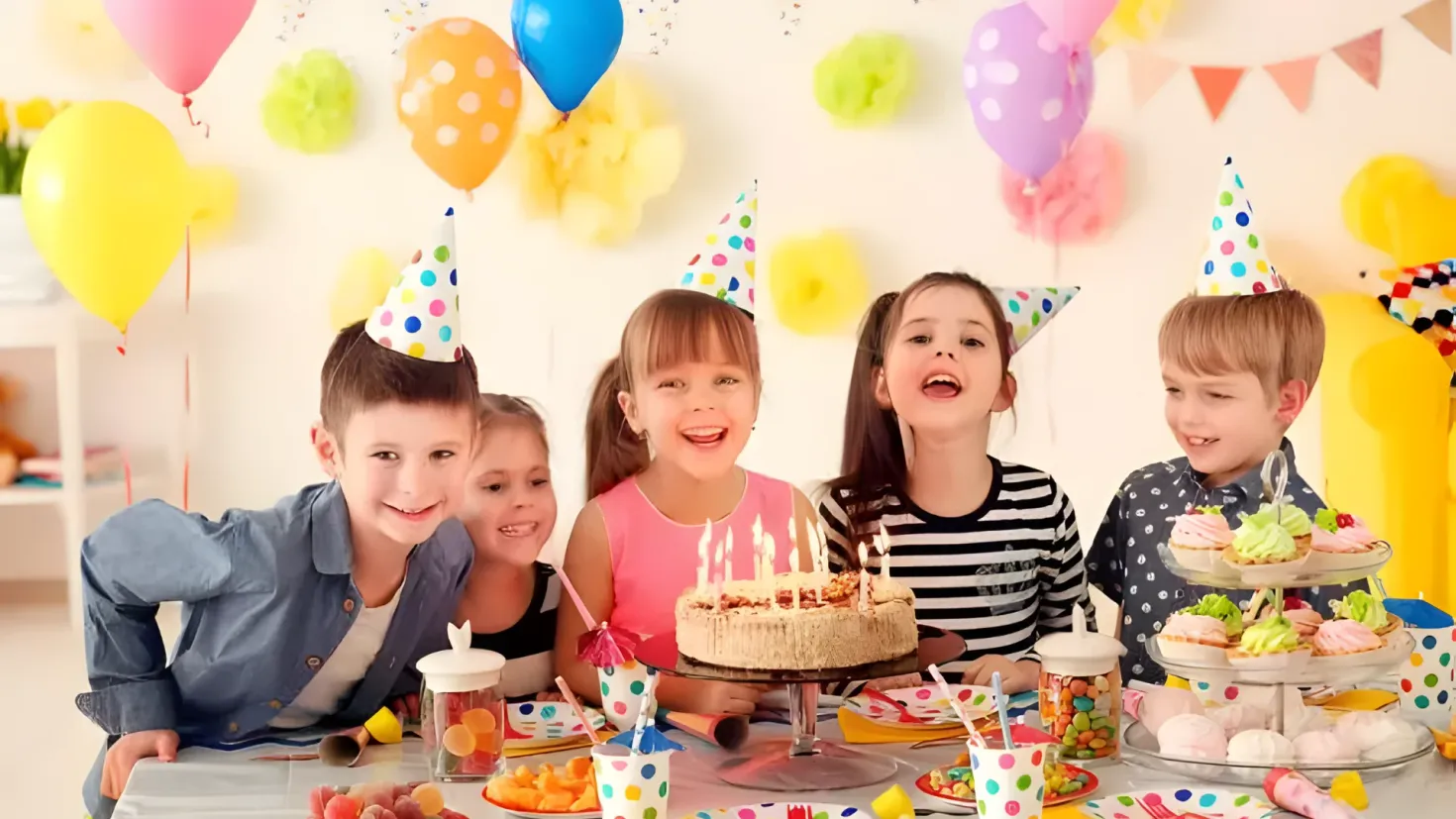 A group of children sitting around a table with a birthday cake adorned with lit candles. On the table there are plates, bowls, spoons, forks, knives, and other food items. In the background, there are colorful balloons and decorative items on the wall, creating a festive atmosphere.