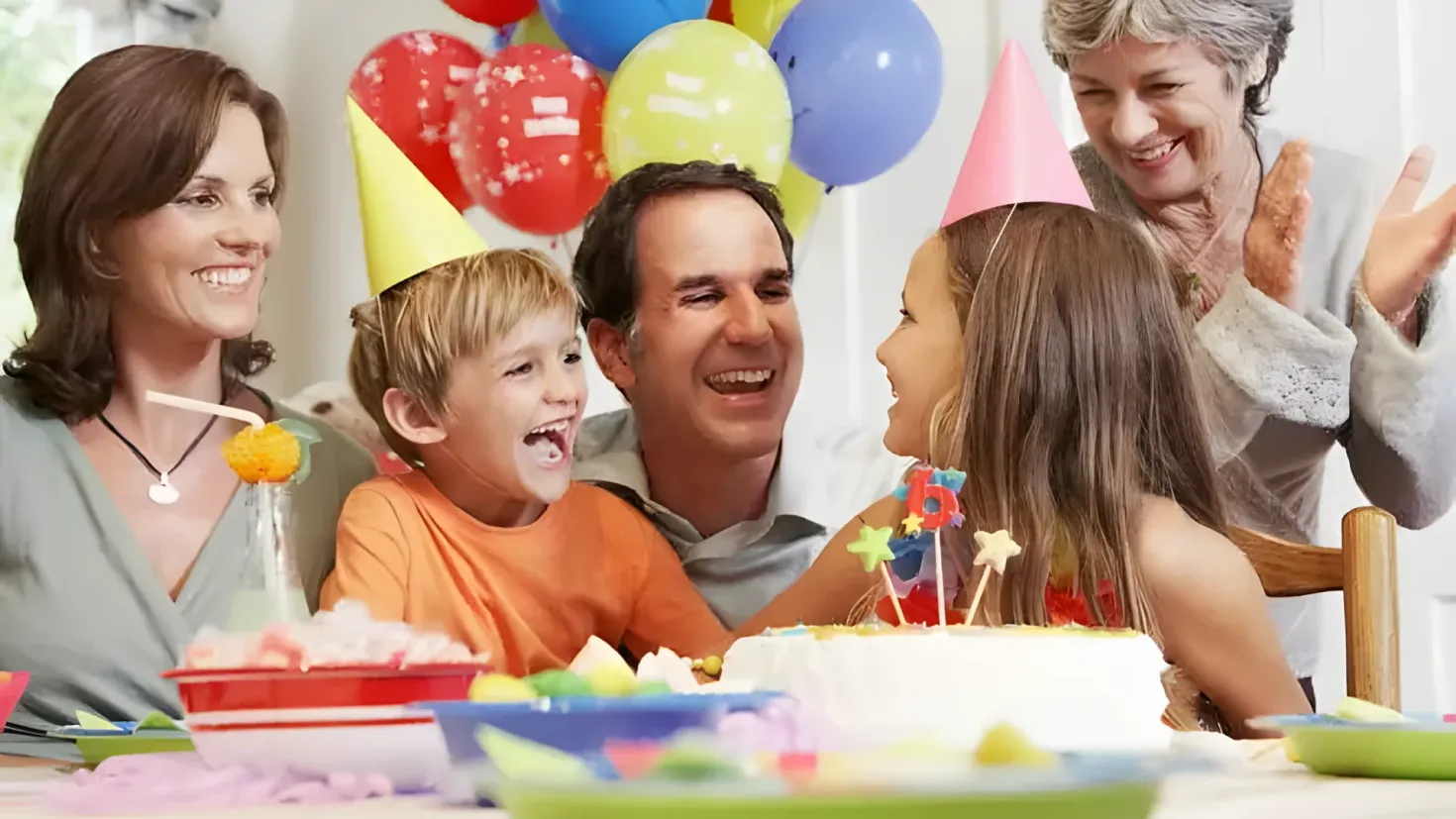 A family gathered around a table with a birthday cake, surrounded by colorful balloons. On the table there are bowls, plates, and other objects, and in the background there is a wall with a window. Everyone is smiling, suggesting that they are celebrating a special occasion.