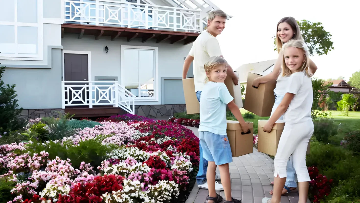 A family of four standing in front of a house, each holding a cardboard box. The house is surrounded by lush greenery, with trees, plants, and flowers in the foreground. The sky is visible in the background, creating a peaceful atmosphere.