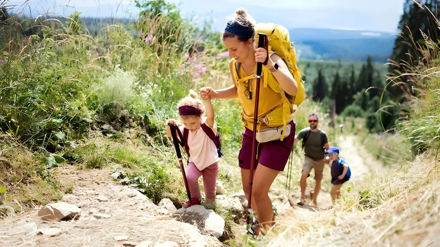 A woman and two children hiking up a trail in the mountains. The woman is wearing a yellow and maroon dress and carrying a yellow backpack, while the two children are wearing different colored dresses. The trail is surrounded by lush green plants and trees, and the sky is a beautiful blue and white.
