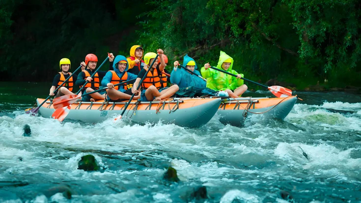 A group of people in a raft on a river, wearing safety jackets and helmets, and holding paddles in their hands. The river is surrounded by lush green trees, creating a peaceful atmosphere.