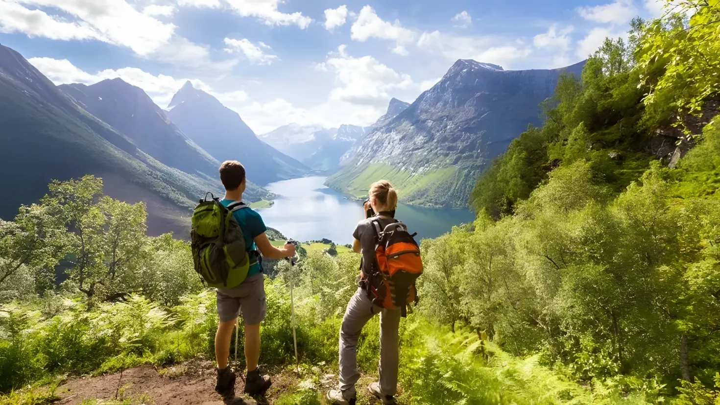 Two people standing on top of a mountain, both wearing backpacks and looking out over a lake surrounded by lush green trees and plants. The sky is filled with white, fluffy clouds, creating a peaceful atmosphere.