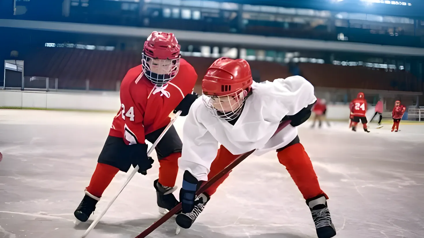 A group of young people playing hockey on an ice rink. They are wearing helmets, gloves, and sneakers, and each of them is holding a hockey stick. In the background, there is a building with lights illuminating the rink.