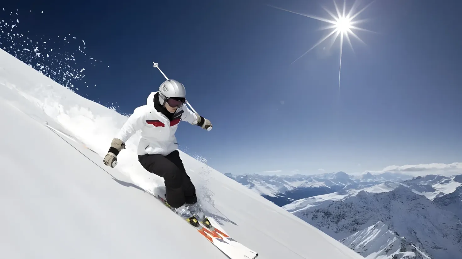 A person skiing down a snowy hill with mountains in the background. They are wearing a white jacket, black pants, a helmet, and gloves, and are holding ski poles in their hands. The sky is a bright blue with the sun shining down.