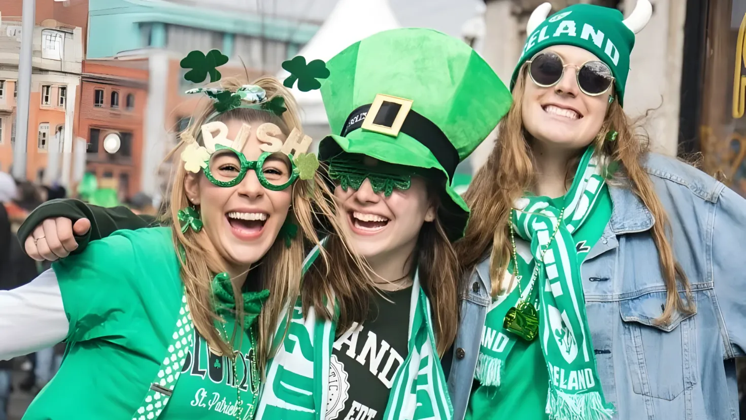 Three women in green hats and scarves posing for a photo in front of a crowd. They are all wearing glasses and hats, and the background of the image is filled with buildings, poles, and a clear blue sky.
