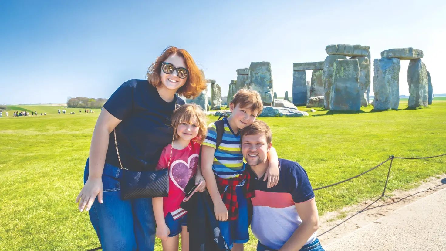 A family of four posing for a photo in front of Stonehenge in Wiltshire, England. The woman is wearing goggles and carrying a bag, while the two children are standing beside her. The background of the image is filled with lush green grass, rocks, trees, and a clear blue sky.