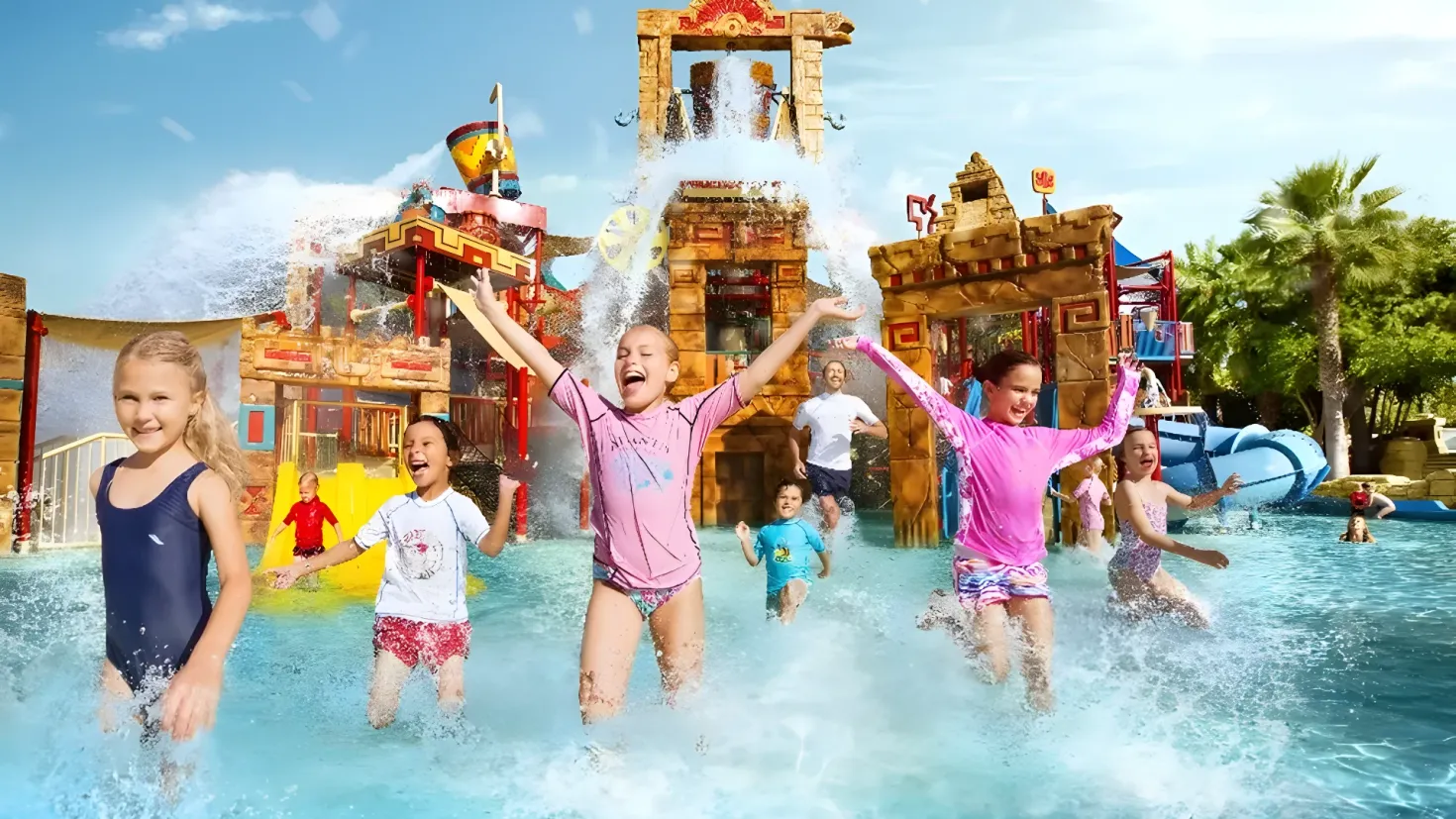 A group of children playing in the water at a water park, surrounded by lush green trees and a bright blue and white sky. In the background, there is a water slide and a few people enjoying the water park.