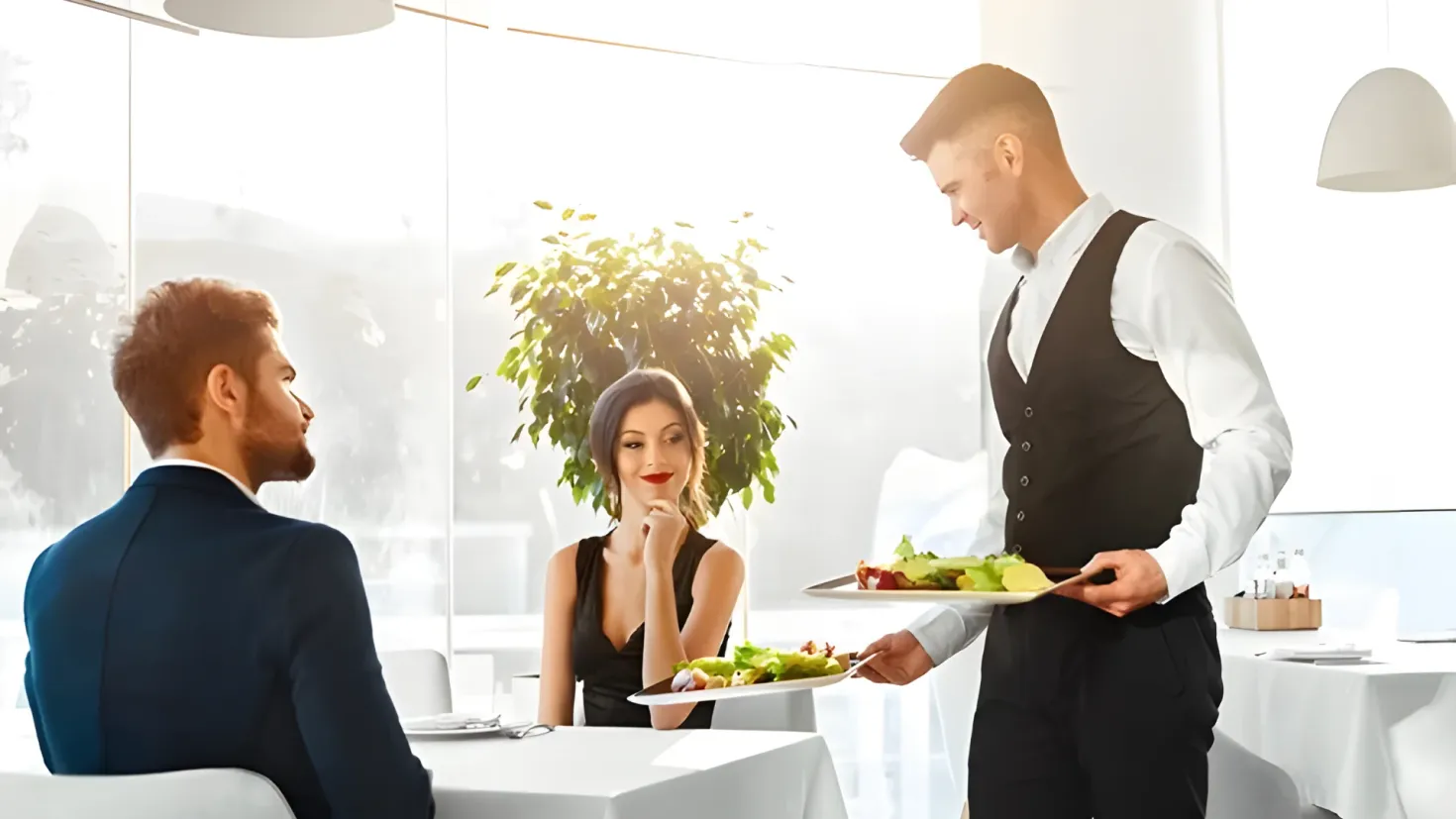A waiter serving food to a couple in a restaurant. The waiter is standing and holding a plate of food, while the couple is seated in chairs around a table with plates, glasses, and other objects. There is a plant in the background and lights hanging from the ceiling.